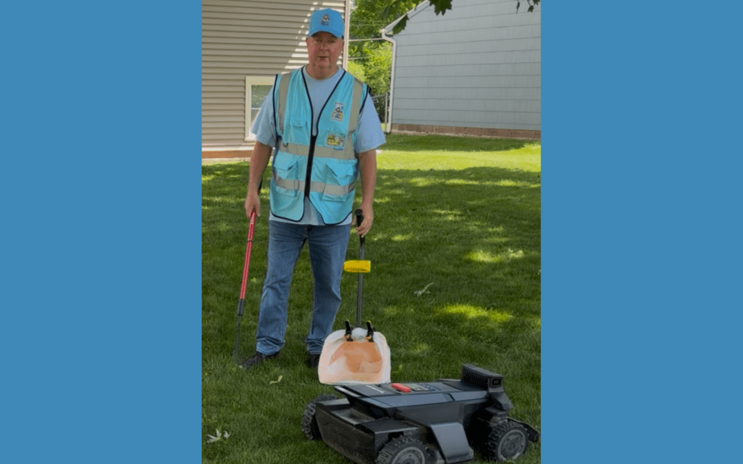 Poopy Monster owner Dan Powell stands in a customer's backyard next to a robotic lawn mower while holding pet waste removal tools, explaining why robot mowers still need a clean yard before mowing.